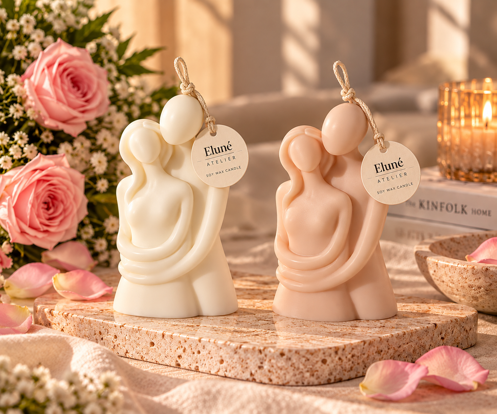 Two sculptural candle holders shaped as embracing figures, one in warm ivory and one in pale peach, displayed on a stone slab with pink roses, petals, and a lit candle in the background. Each figure has a hanging tag reading 'Elune Atelier'.