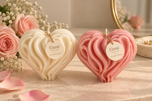 Two heart-shaped wax candles with textured swirls, one cream and one dusty pink, displayed on a marble surface with pink rose petals and flowers. Each candle has a hanging tag reading 'Eluné Scents'.