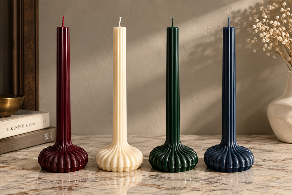 Four ribbed taper candles in burgundy, cream, forest green, and navy blue, displayed on a marble surface against a textured beige wall.
