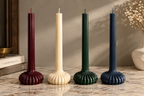 Four ribbed taper candles in burgundy, cream, forest green, and navy blue, displayed on a marble surface against a textured beige wall.